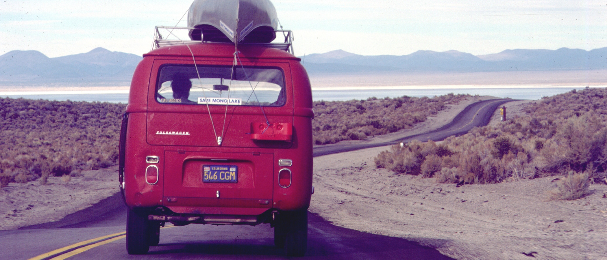 The back of a red bus with a canoe on the roof and a Mono Lake Sticker drives down the road towards the lake.