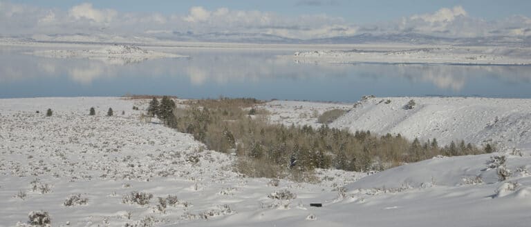 The islands on Mono Lake and surrounding mountains are covered in snow, and the still water reflects the light blue and cloudy sky above.