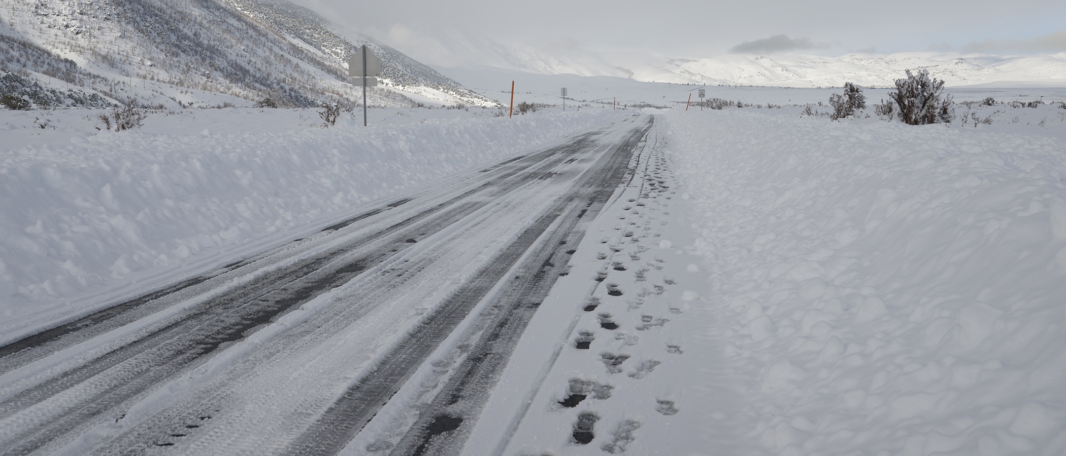 A road is barely visible between huge piles of plowed snow, and stretches into a completely white mountainous landscape.