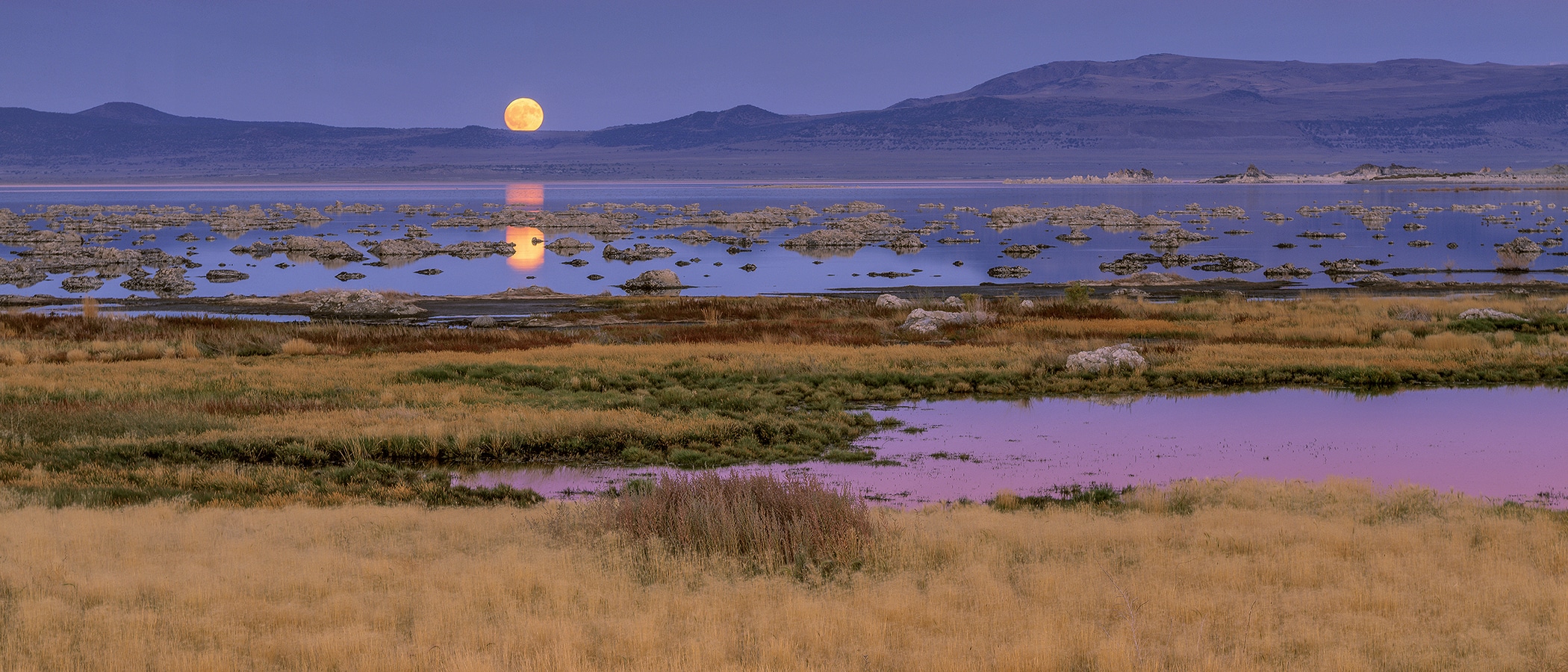 A huge orange full moon hangs in a purple sky and is reflected over the blue-purple water of Mono Lake.