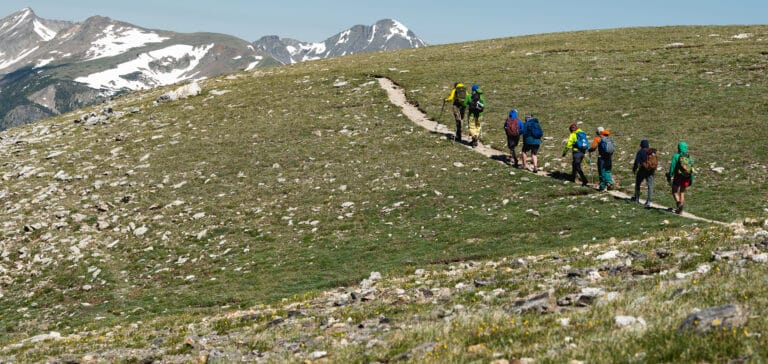 8 hikers with colorful packs and poles walk along a dirt narrow path on a grassy hill towards snowcapped mountains.