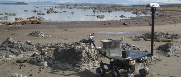 The mars rover, a metal machine with six wheels sits on the beach of Mono Lake near outcroppings of tufa.