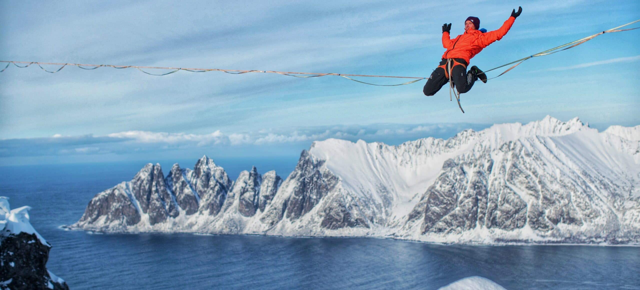 A man in an orange jacket sits on a tight rope with his hands outstretched. Huge snowy mountains and blue water loom below the high wire.