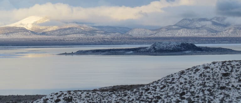 An island in Mono Lake is covered in snow, and so are the hilly mountains behind it. The sky is cloudy with yellow sunlight shining through onto the glassy light blue surface of the water.