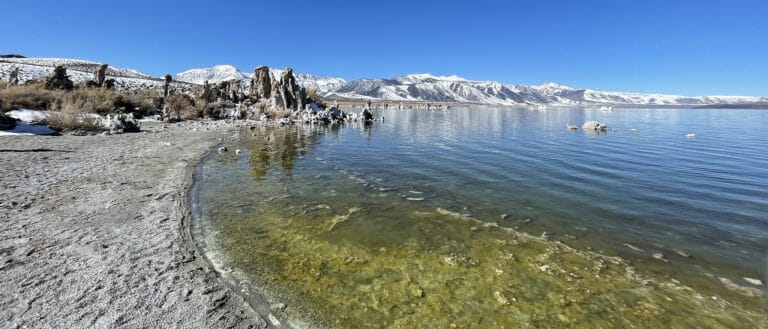 Looking along a sandy beach, to the right a lake, in the distance tufa formations and snow capped mountains under a clear blue sky.