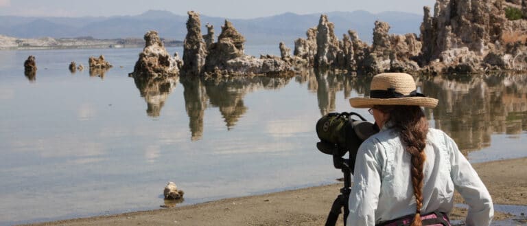 A woman on the shore of Mono Lake looking through a spotting scope out over the water with tufa towers in the background.