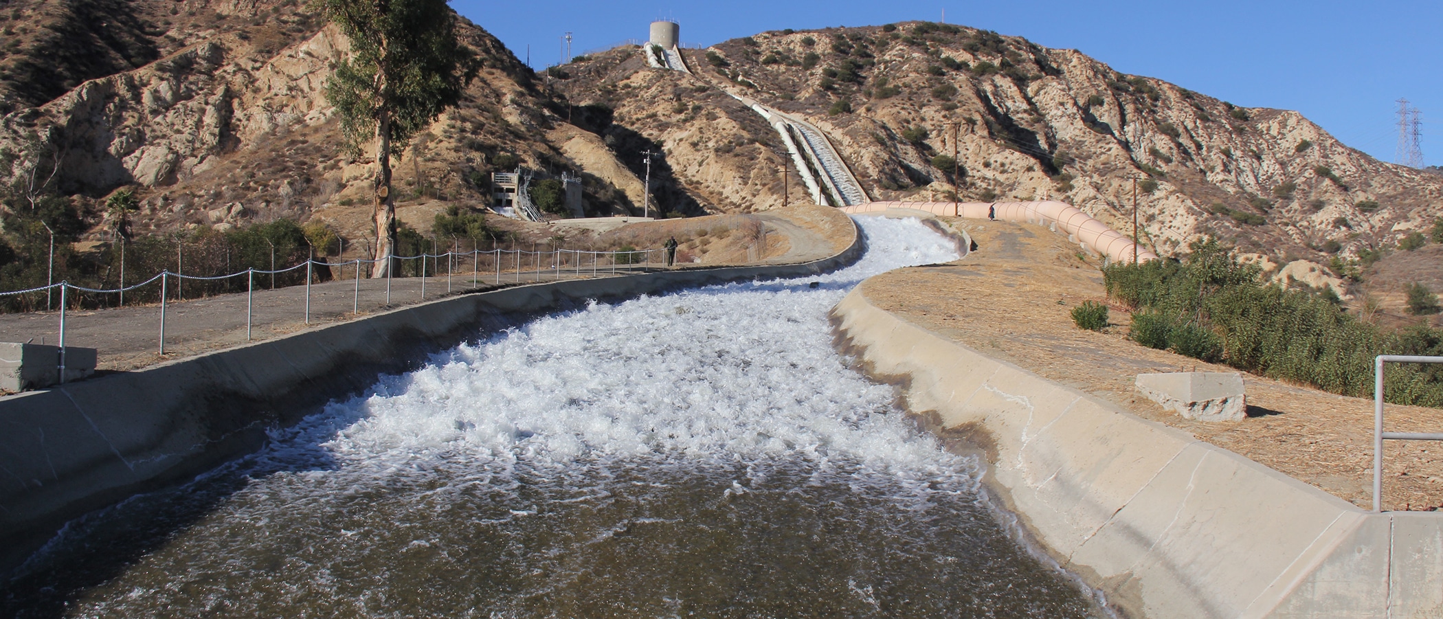 White water rushes down a cement trench from the top of a rocky brown hill.