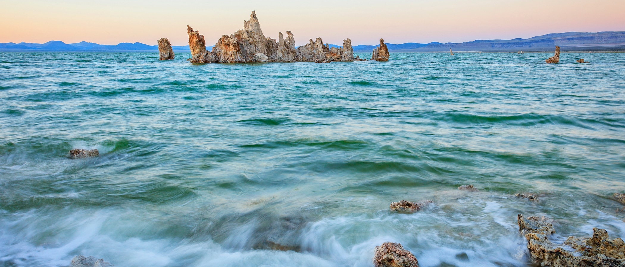 Turquoise Mono Lake water surface is rough with small waves, and tufa towers rise up in front of blue mountains and a pink and orange glowing sky.