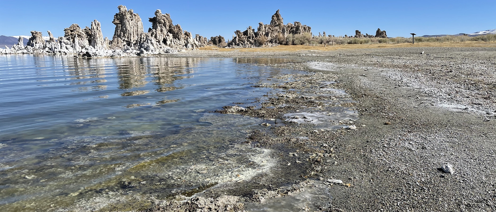 Mono Lake water reflects the blue sky, and water it frozen in bumpy puddles on top of the tufa beach. Tufa towers loom in the background.