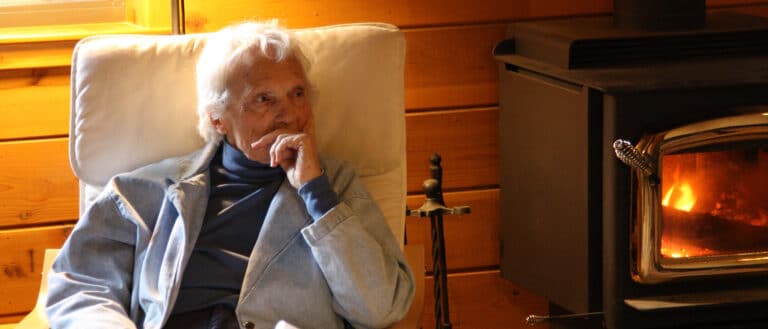 A white haired woman sits in a chair next to a fireplace.