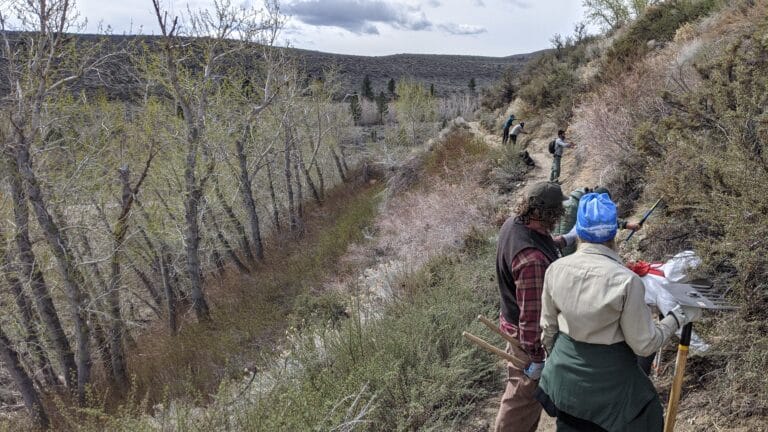 Two trail workers converse on a thin dirt trail between sagebrush. Other workers can be see down the trail.