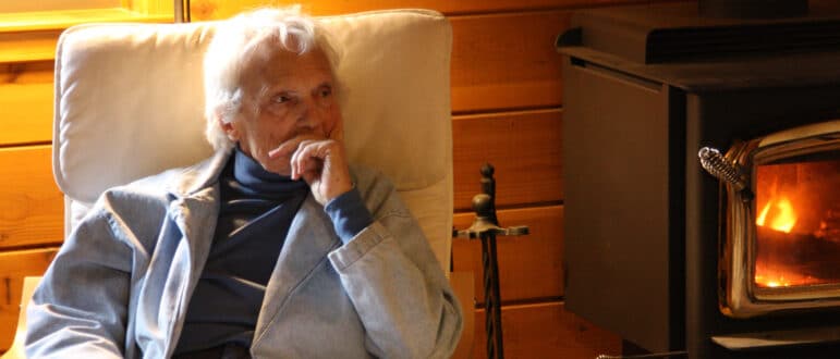 A white haired woman sits in a chair next to a fireplace.