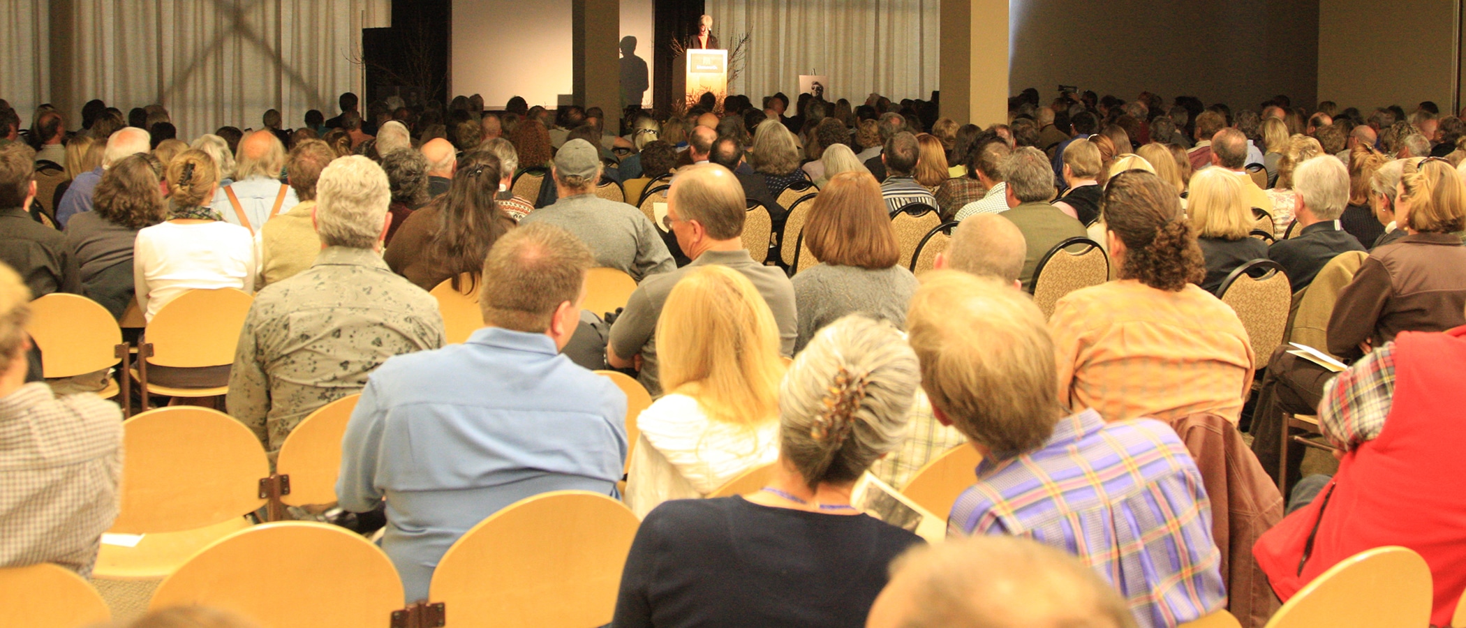 A crowd of people seated in chairs looks toward a stage where someone stands at a podium.