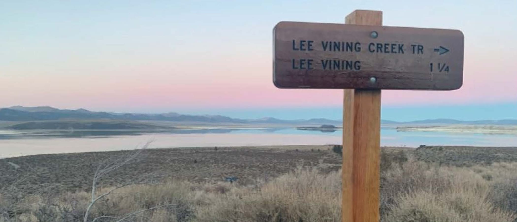 A wooden trailhead for Lee Vining Creek stands against a sunset gradient of blur, yellow, and pink reflected in the Mono Lake water.