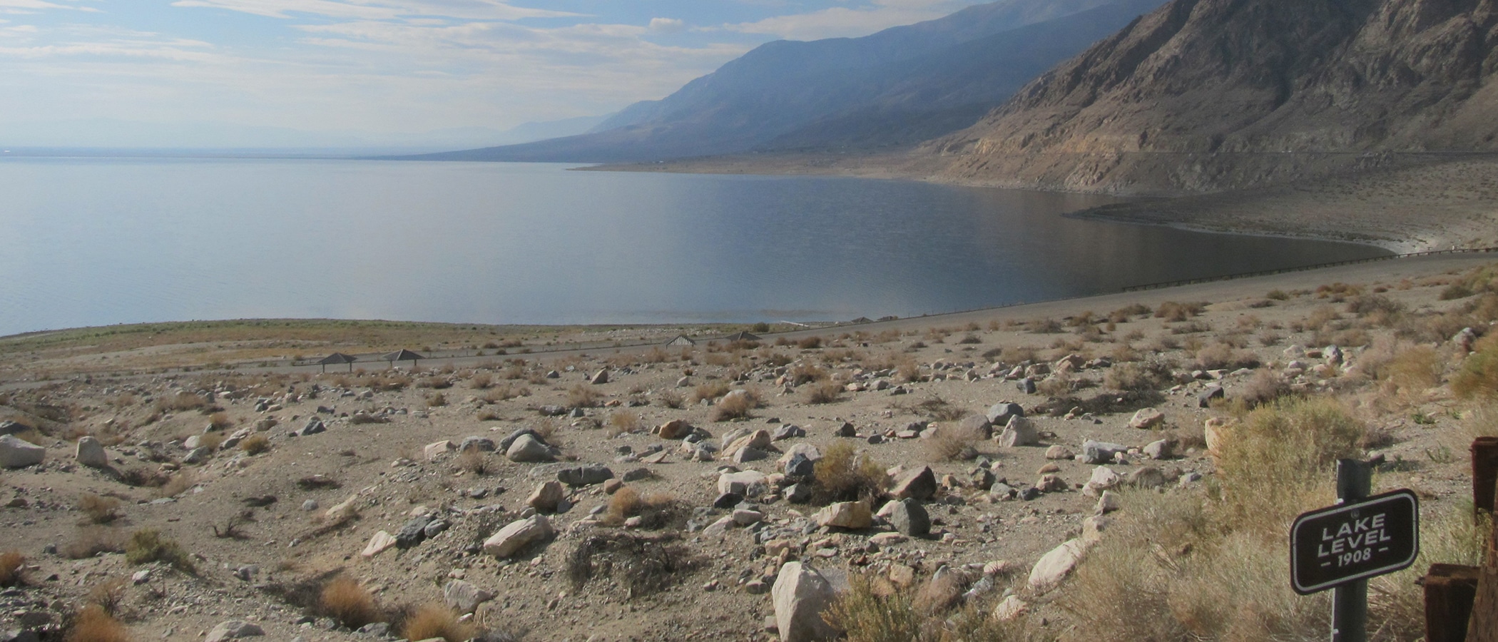 A glassy lake reflects a cloudy and blue sky, with a sandy and rocky beach and a sign marking the lake level.