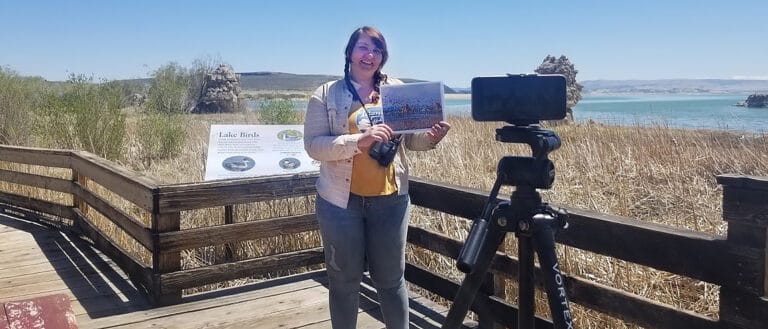 A person stands on a wooden boardwalk holding a laptop in front of a tripod holding a cell phone, in the background is a marsh, lakeshore, and blue skies.