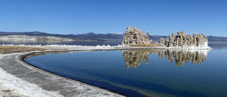 A white and grey shoreline wraps around to tufa towers perfectly reflected in a bright blue Mono Lake with clear blue skies above.