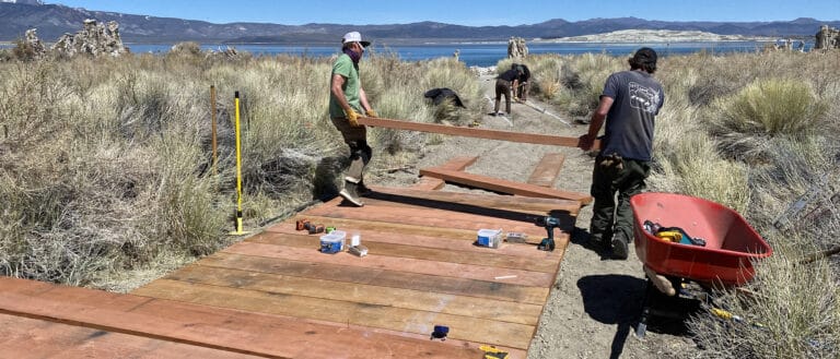 People carrying boards into place on a path toward Mono Lake, mountains are on the horizon under a clear blue sky.