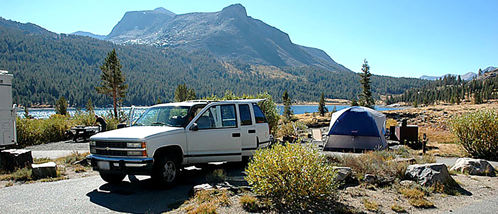 A tent setup in a campsite, a white SUV is parked nearby, a lake and mountain are in the background.