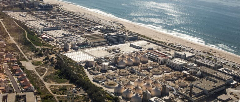 Aerial view of a wastewater treatment plant just inland of a beach along the ocean, the sun reflects off the ocean.