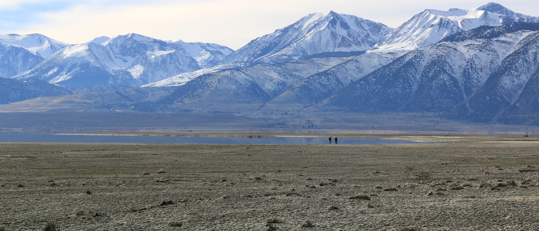 Two people stand along the shore of Mono Lake with snow capped mountains large in the frame behind them.