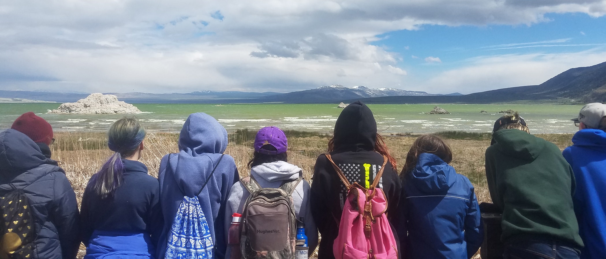 Eight students stand in a line looking out at a very green Mono Lake with waves on it and with a tufa island and the Mono Craters in the distance, and they are all wearing cool-weather outdoor gear.