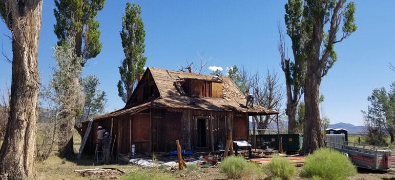 People working on an old farm house flanked by green leaved trees and a blue sky.