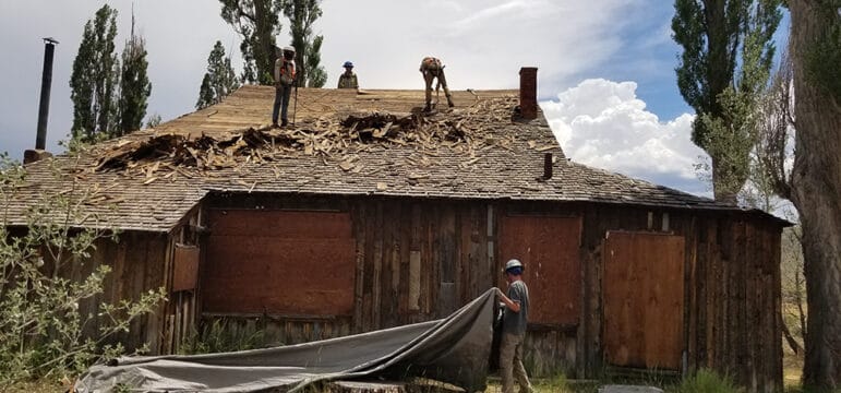 People working to put a tarp on an old farm house flanked by green leaved trees and a cloudy sky.