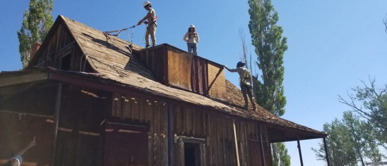 People working on the roof of an old decaying farm house under a clear blue sky.