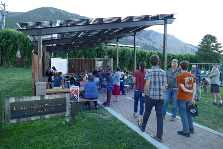 A crowd filtering in under an outdoor structure surrounded by a grassy park.