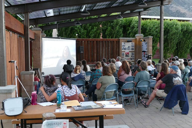 A seated crowd watching a film under an open walled roof.