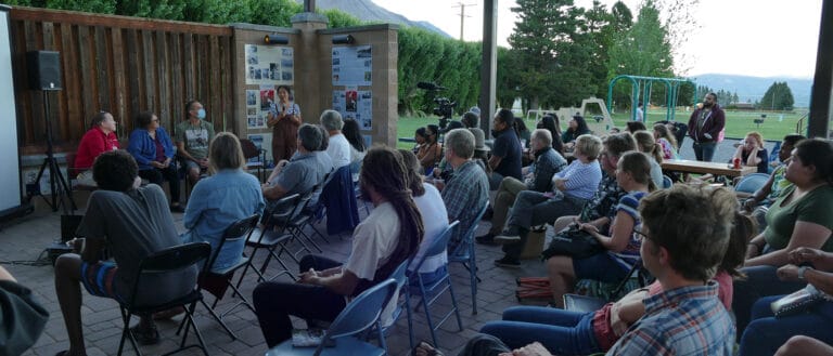 A crowd seated outdoors, a panel stands in front of them with a microphone.