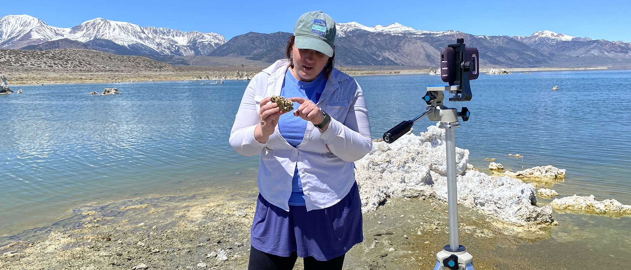 A person stands in front of a cell phone on a tripod with their back toward Mono Lake holding a piece of tufa and pointing at it, the Sierra Nevada is snow capped in the distance against a blue sky.