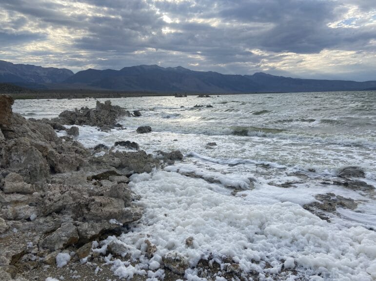 Waves crashing against tufa and a thick layer of foam on the shore of Mono Lake with mountains in the background, the sky is overcast.