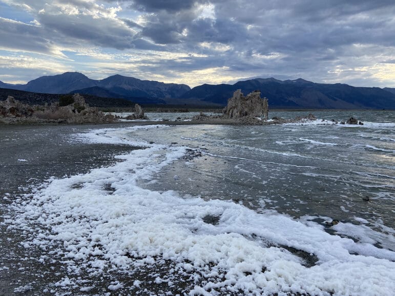 Waves crashing against tufa and a thick layer of foam on the shore of Mono Lake with mountains in the background, the sky is overcast.