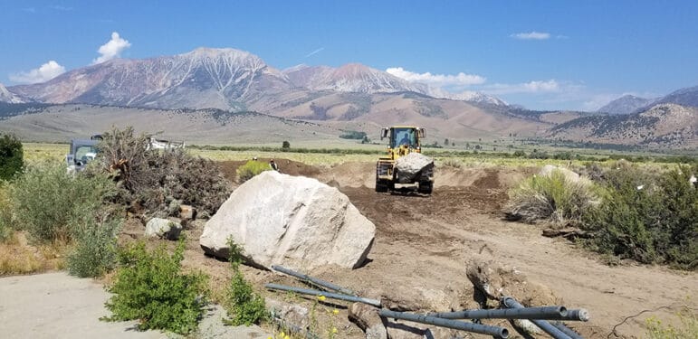 A front end loader moves boulders around on bare dirt, the mountains and a few clouds in a clear blue sky are in the distance.