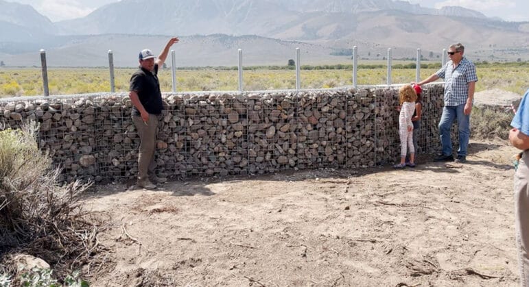 People standing in front of a gabion fence with brush and mountains in the background.