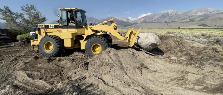 Front end loader moving a boulder with sagebrush and the eastern flank of the Sierra Nevada in the background.