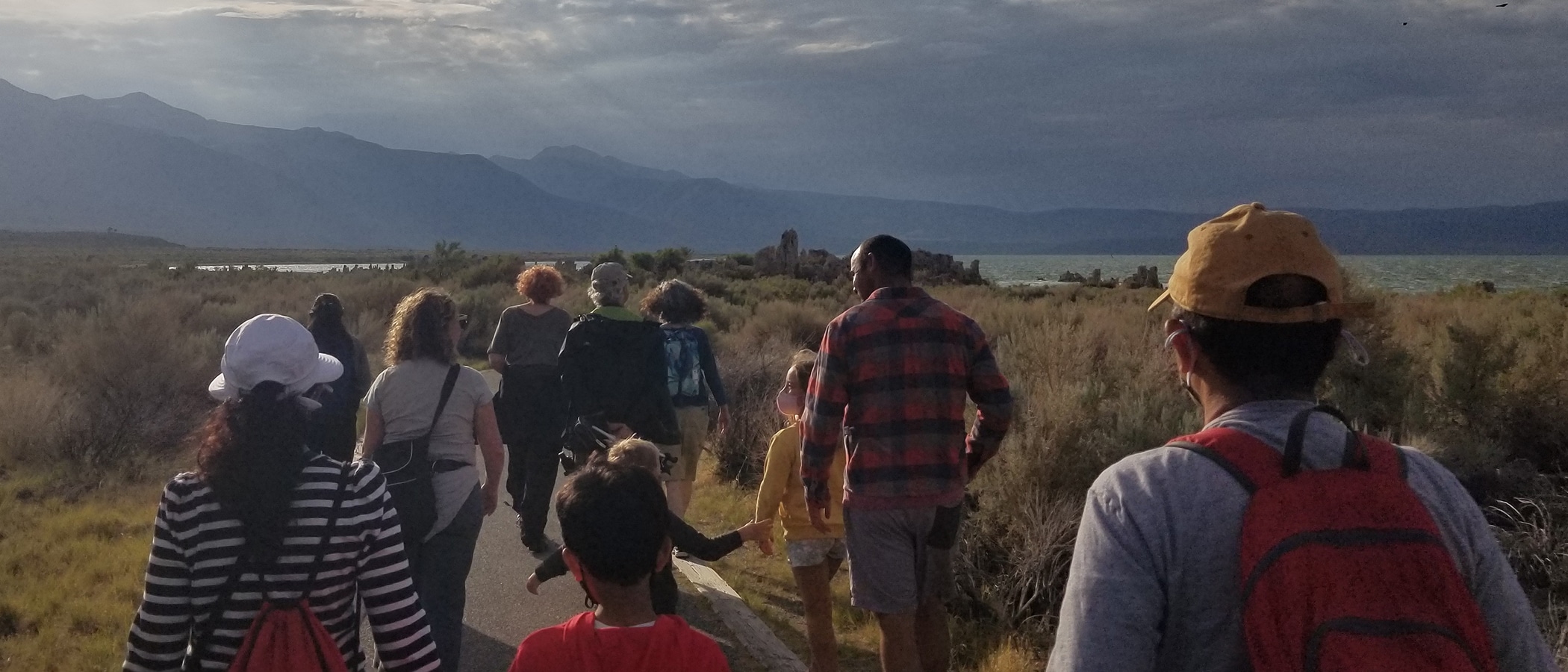 People walking down a path toward tufa, Mono Lake and the mountains are in the distance, the sky is cloudy.