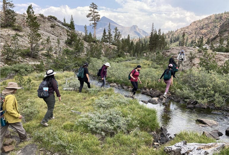 People crossing a small stream in a high country meadow with trees and mountain ridges in the background.