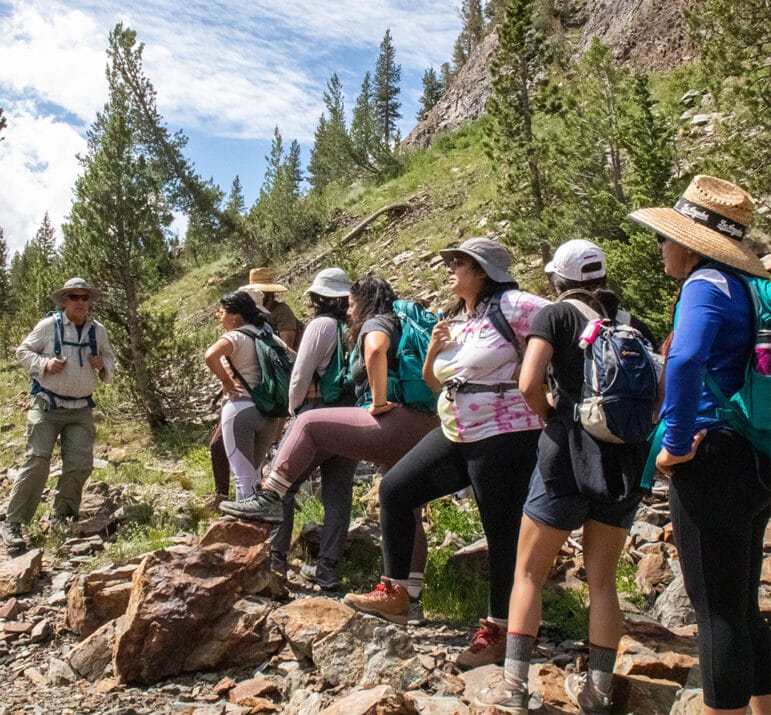 A line of people stand wearing packs along a rocky trail along the slope of a hill with small pine trees on it.