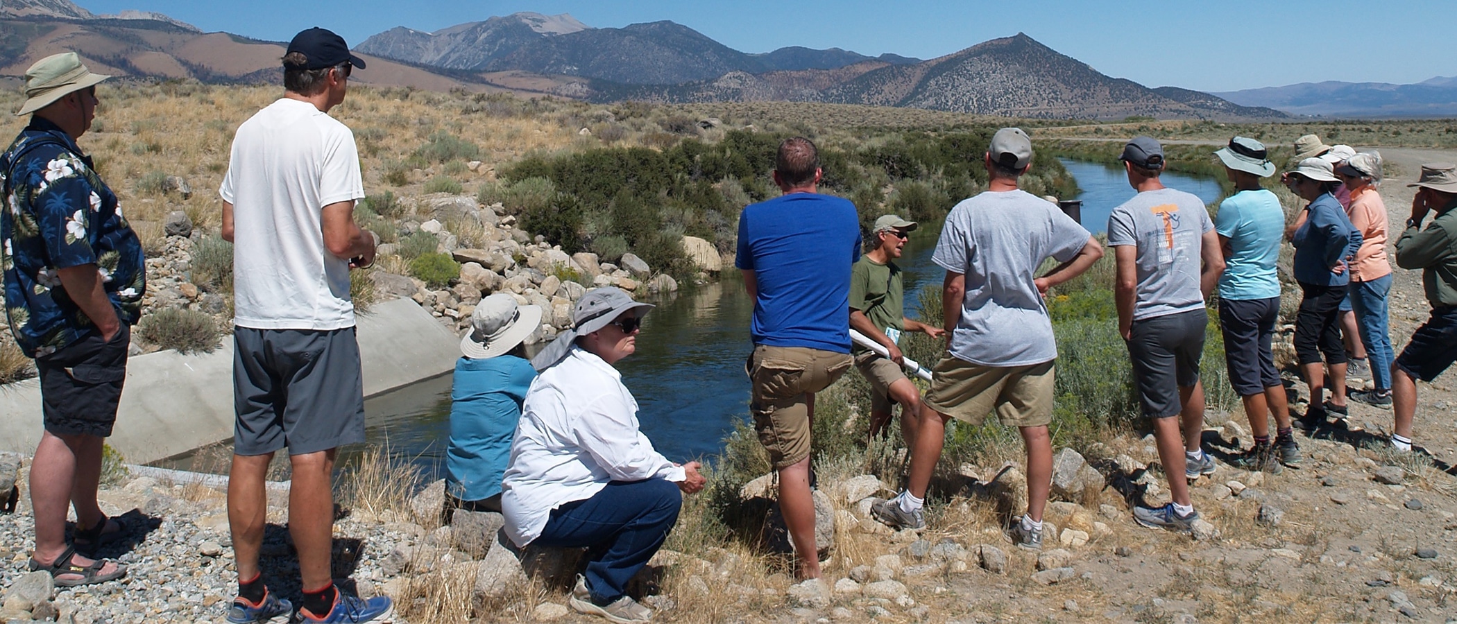 A dozen people stand facing an aquaduct full of water and a person with their back to the watercourse gesturing, around the aquaduct path is desert scrub and mountains in the distance under a blue sky.