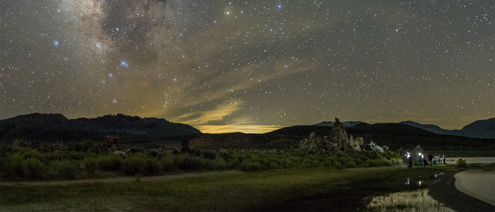 The milky way and stars are seen in the sky, hills form dark silhouettes beneath the sky, tufa formations and people holding lights are visible in the foreground.