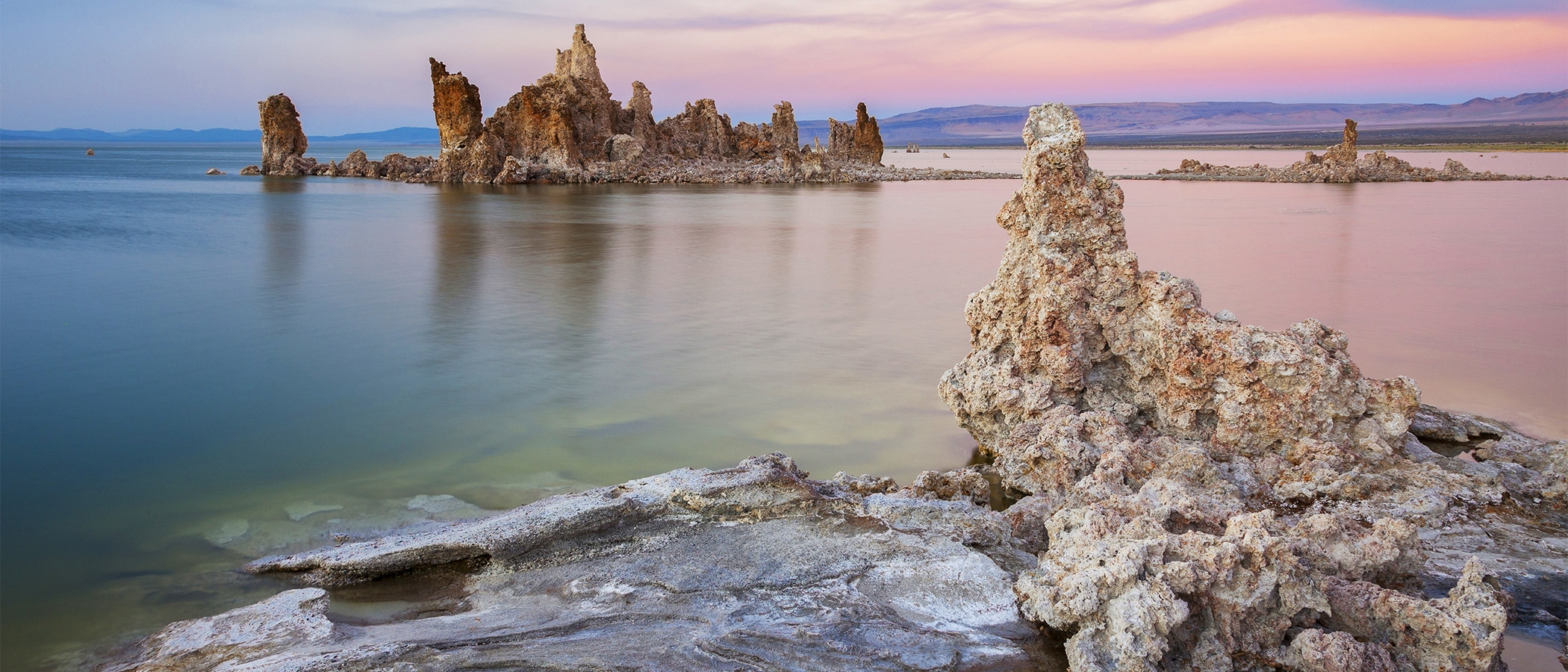 A lake surface with tufa formations, the water is reflecting intense pink and purple colors in the cloudy sky.