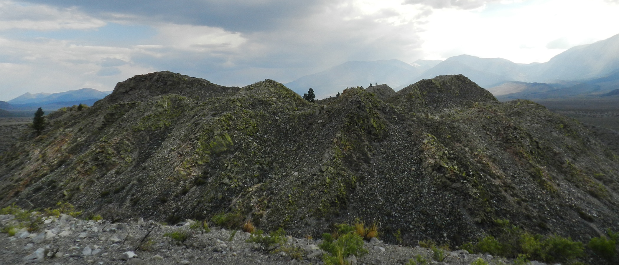 A rugged hill of black jumbled rock dotted by scattered green bushes under a cloudy sky.