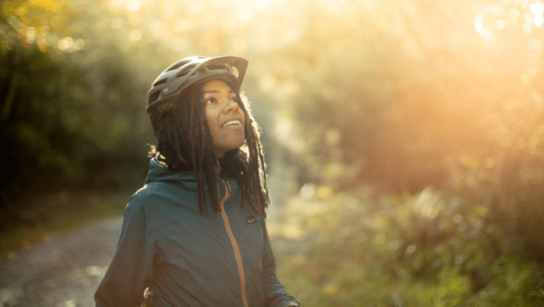 A person with a bike helmet stands looking upward, greenery and the glow of light fills the background.