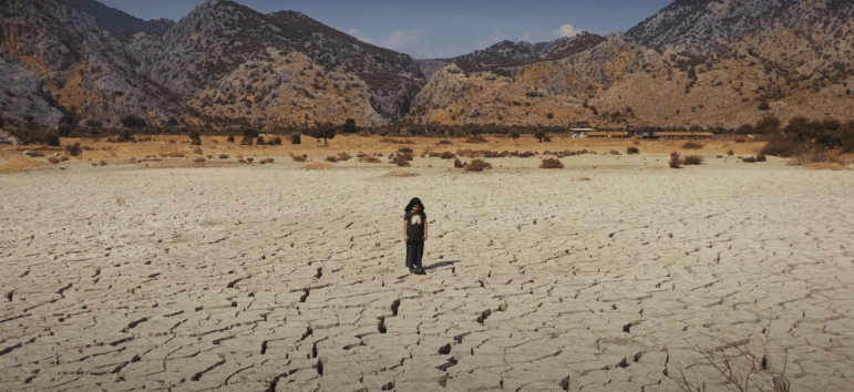 A person stands in a flat area of dry cracked soil with arid looking mountains in the background.