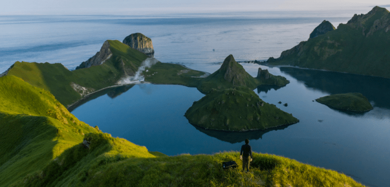 Water surrounds sharp volcanic looking green covered hills, a person stands in the foreground on top of one of the hills.