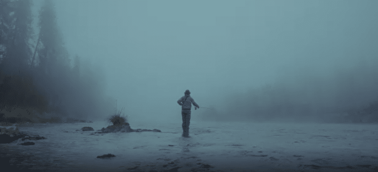 A person stands in shallow water holding a fishing pole surrounded by fog, faint shapes of conifer trees can be seen on the banks.