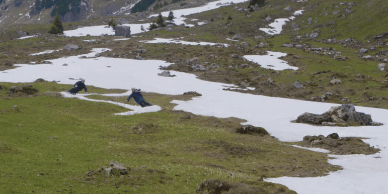 Two people are mid turn skiing down a slope with patchy snow and lots of grass and rocks.